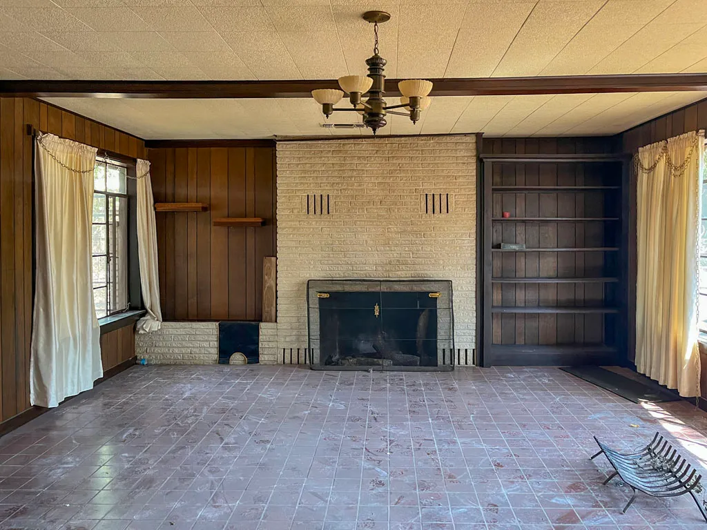 Outdated 1970s living room with wood paneling, brick fireplace, and tile flooring before kitchen conversion in San Antonio TX